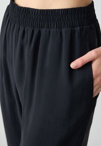 Pantalons noirs légers avec une taille élastique et des poches latérales. Texture lisse avec une coupe décontractée ; le tissu tombe doucement.