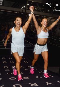 Twee vrouwelijke atleten in witte sportkleding en roze schoenen vieren door hun gesloten handen boven hun hoofd te heffen op een donkere binnenbaan.