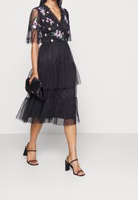 Robe en tulle noire avec broderie florale, encolure à volants et silhouette à étages. Portez-la avec des sandales noires à talons et une pochette noire.