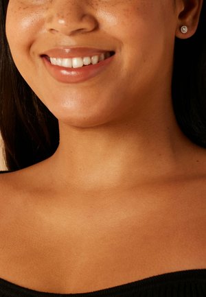 Close-up van de hals en het glimlachende gezicht van een persoon, met een gladde huid, een warme ondertoon en fonkelende oorknopjes. Zwart shirt zichtbaar.