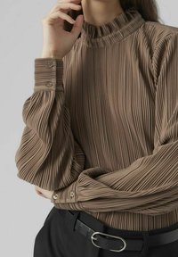 Blouse marron plissée avec un col haut froncé et de longues manches bouffantes. Dotée de poignets boutonnés et d'un tissu texturé. Portée avec une ceinture noire.