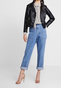 Femme portant une veste en cuir noire, un chemisier blanc à motifs, un jean bleu taille haute avec revers, et des sandales à talons nus, debout sur fond blanc.