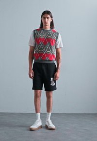Graue Strickweste mit roten Rautenmuster über einem weißen T-Shirt, kombiniert mit schwarzen Shorts mit Grafik und weißen Sneakers.