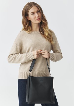 Black leather handbag with a structured shape and adjustable strap, featuring silver hardware and a textured finish, held by a person in a beige sweater.