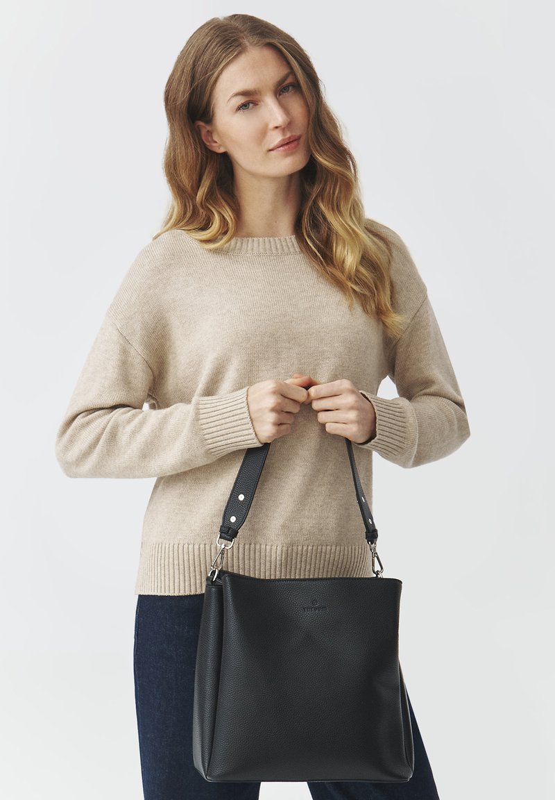 Borsa a mano in pelle nera con forma strutturata e tracolla regolabile, dotata di accessori in argento e finitura testurizzata, tenuta da una persona in un maglione beige.