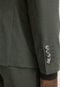 Giacca a giacca verde che mostra un tessuto strutturato, un design aderente e cinque bottoni metallici sul polsino con dettagli di cuciture visibili.