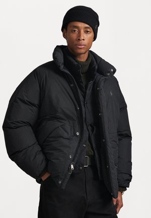 Junger Mann trägt eine schwarze Mütze und eine schwarze Steppjacke über einem dunkelgrünen Rollkragenpullover und schwarzen Hosen vor einem schlichten weißen Hintergrund.