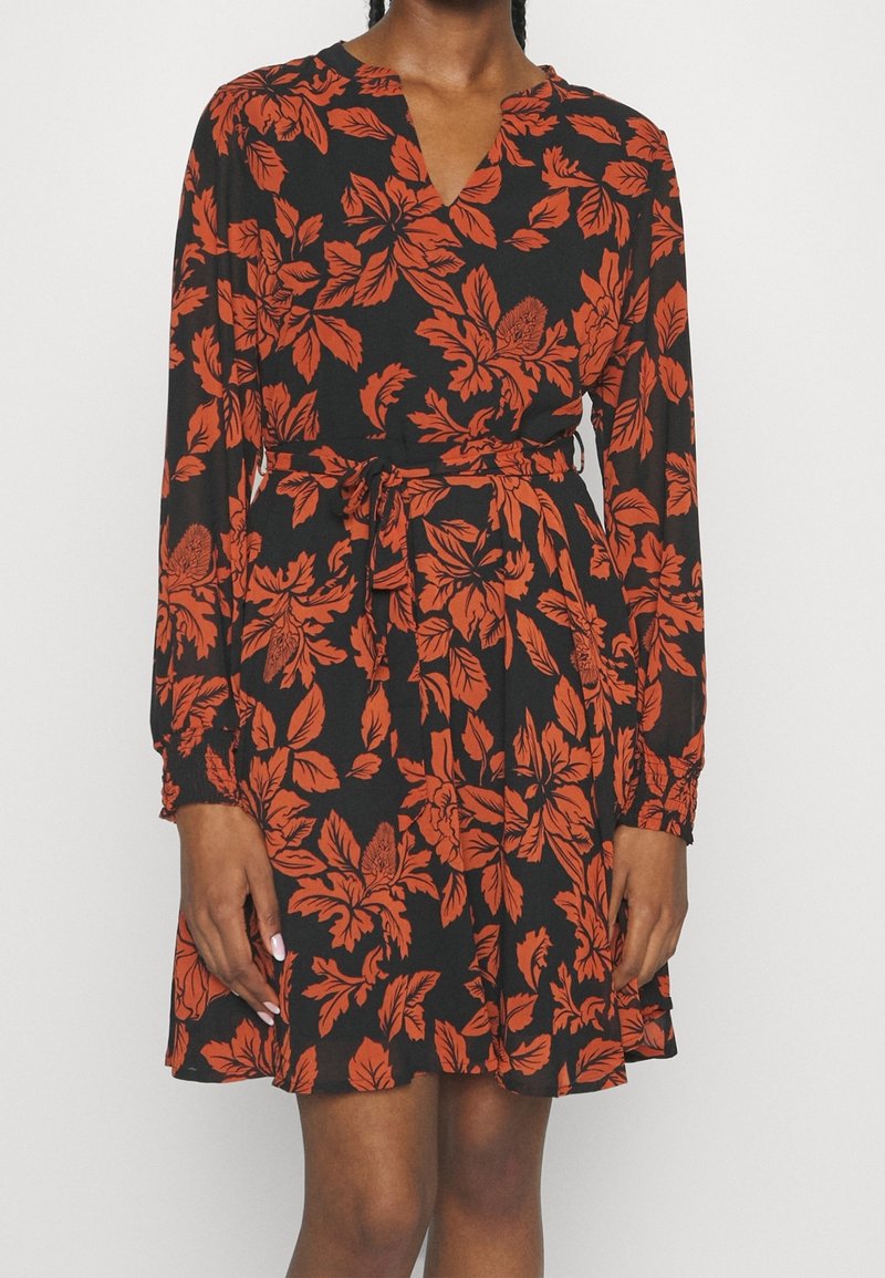 Robe noire à manches longues avec motif floral orange, encolure en V et ceinture nouée à la taille, portée par une personne debout.
