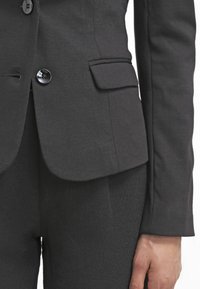 Gros plan sur une personne portant une veste de costume sombre sur mesure avec des boutons et un pantalon assorti, montrant le bras droit et la main détendus.