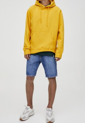 Personne portant un sweat à capuche jaune, un short en jean bleu et des baskets blanches, debout devant un fond uni.