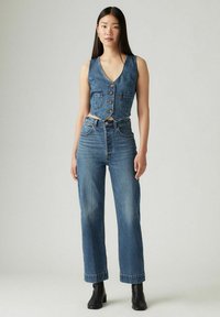 Gilet di denim con scollo a V e bottoni, abbinato a jeans a gamba dritta a vita alta in blu medio. Tessuto texturizzato con due tasche frontali.