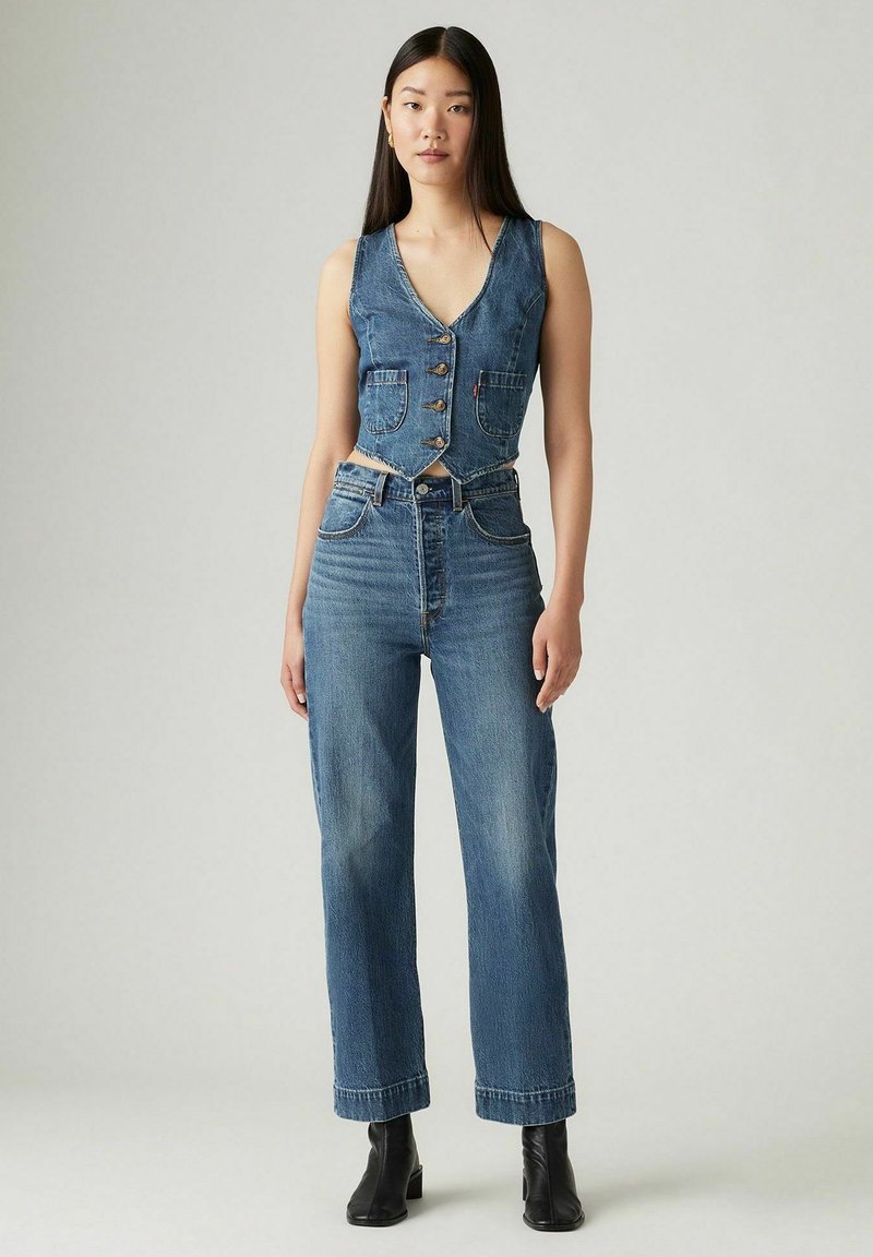 Gilet di denim con scollo a V e bottoni, abbinato a jeans a gamba dritta a vita alta in blu medio. Tessuto texturizzato con due tasche frontali.