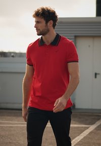 Rotes Poloshirt mit schwarzen Kragenakzenten, kurzen Ärmeln und einem dezenten Logo. Trägt schwarze Hosen, Modell läuft auf einem Dach.