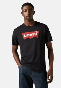 Joven con una camiseta negra de Levi's con un logo rojo y blanco, combinada con jeans azules, de pie frente a un fondo claro y liso.