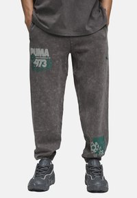 Grå joggerbukser i bomuldsblanding med elastiske manchetter. Har et teal grafisk "PUMA" logo og blomsterdesign på venstre ben.