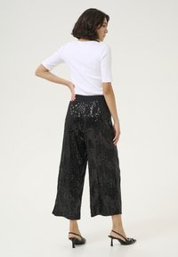 Pantaloni neri a gamba larga con texture di paillettes, dotati di una vita nera. Abbinati a un top bianco aderente, il modello è in piedi con la schiena rivolta.