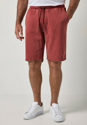 Mand iført rustfarvede shorts med snøre og hvide sneakers står med den ene hånd i lommen mod en ensfarvet baggrund.