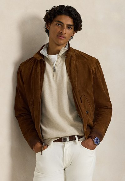 Jonge man met krullend haar, draagt een bruine suède jas over een beige trui met een kwart rits en witte broek, handen in de zakken, neutrale achtergrond.