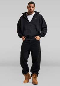 Sort zip-up hoodie med en kængurulomme, parret med løse sorte bukser. Tan støvler fuldender looket. Minimalistisk branding på hoodien.