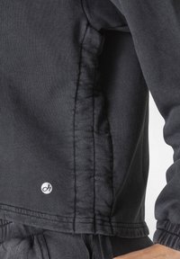 Chaqueta gris oscuro hecha de un tejido suave y texturizado, con cierre de cremallera y un pequeño logo blanco visible cerca del dobladillo. Presenta costura fruncida.