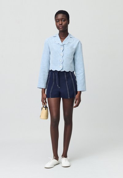 Giacca corta azzurra con orlo ondulato e bottoni, abbinata a pantaloni corti blu navy a vita alta con cuciture bianche e una piccola borsa intrecciata.