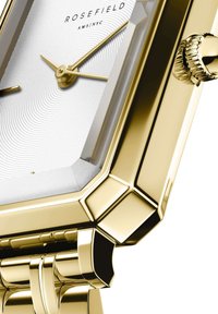 Reloj rectangular con tono dorado y esfera plateada texturizada, que presenta manecillas pulidas, una corona estriada y una pulsera lisa.