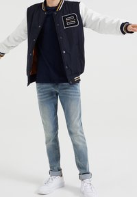 Person som bär en marinblå och vit varsityjacka med "B Bad Boys"-patch, mörkt skjorta, uppvikta ljusblå jeans och vita Puma-sneakers.