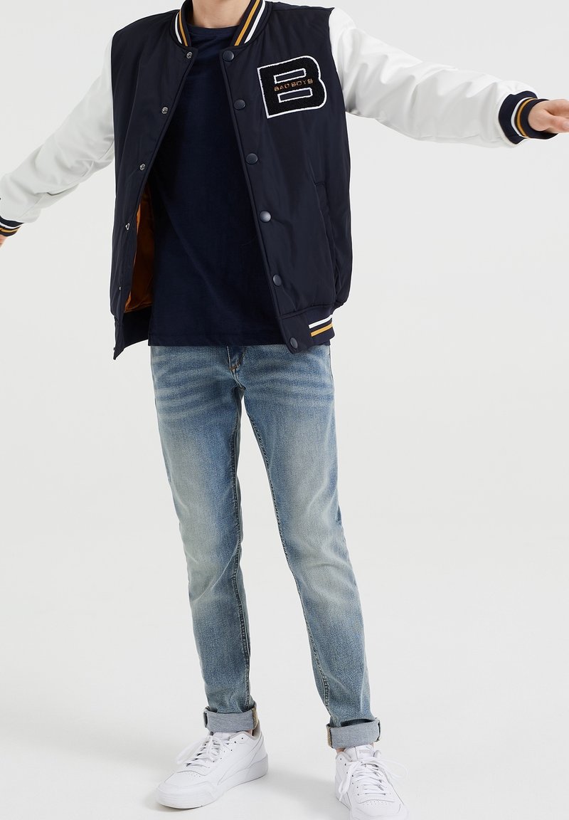 Person som bär en marinblå och vit varsityjacka med "B Bad Boys"-patch, mörkt skjorta, uppvikta ljusblå jeans och vita Puma-sneakers.