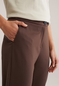 Pantaloni maro croiți, cu textură netedă, design simplificat și închidere cu un singur nasture la talie. Mână plasată în buzunar.