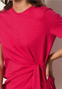 T-shirt fuchsia à manches courtes en tissu lisse, avec un détail noué sur un côté et un col rond classique.