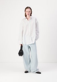 Oversized white striped shirt, light blue wide-leg pants, black flats, and a small black handbag; smooth fabric, relaxed fit, and subtle pattern.