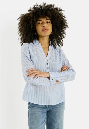 Mujer con cabello rizado y voluminoso, vestida con una camisa de rayas azul claro y jeans, de pie con los brazos cruzados, expresión neutral.