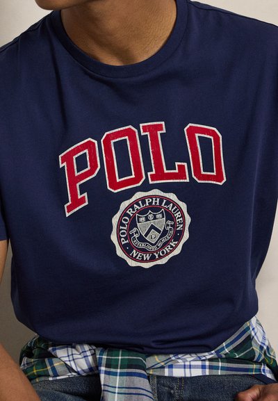 Άνδρας που φοράει μπλε σκούρο t-shirt με μεγάλο κόκκινο κείμενο "POLO" και στρογγυλό λογότυπο Polo Ralph Lauren New York στο στήθος.
