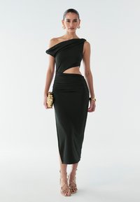 Zwarte, nauwsluitende jurk met asymmetrische halslijn, zijuitsnijding en geplooide textuur. Model houdt een gouden clutch met manchet vast.