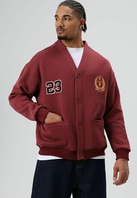 Bordeauxrode cardigan-achtige jas met een V-hals, knopen, voorzakken en geborduurde details waaronder het nummer "23" en een wapen.