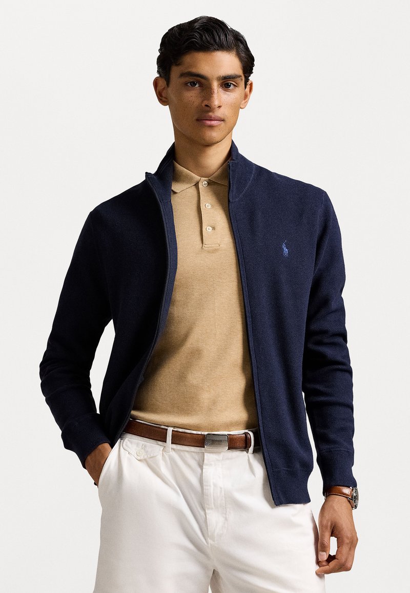 Pulover bleumarin cu fermoar, din tricot cu model texturat, purtat peste un polo bej. Prezintă un guler ușor, mâneci lungi și manșete dublate. Pantaloni albi și o curea maro sunt vizibile.
