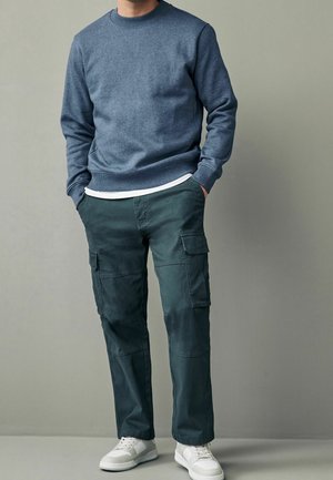 Blaues Baumwoll-Sweatshirt mit Rundhalsausschnitt, kombiniert mit dunklen grünen Cargo-Hosen mit seitlichen Taschen, ergänzt durch weiße Sneaker.