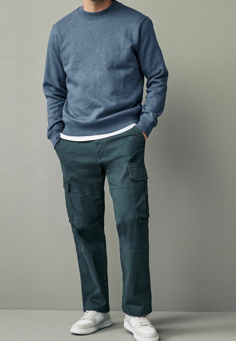 Felpa in cotone blu a girocollo abbinata a pantaloni cargo verde scuro con tasche laterali, completata da sneakers bianche.