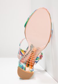 Sandale à bout ouvert avec des sangles en tie-dye coloré, semelle intermédiaire transparente avec motif rayé et semelle beige, inclinée contre une surface blanche.
