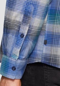 Blauw geruit shirt met variërende tinten blauw, wit en zwart. De stof is glad met een knoopdetail bij de manchetten.