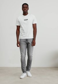 Hvit bomulls T-skjorte med svart logo på brystet, sammen med grå slim-fit jeans og hvite sneakers, på en nøytral bakgrunn.
