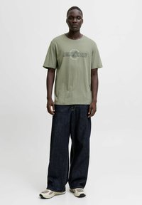 Muž v světle zeleném tričku Jack & Jones, tmavých širokých džínách a béžových teniskách, stojící na pozadí s jednoduchou bílou barvou.