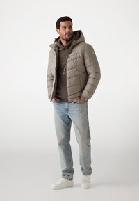 Jachetă căptușită de culoare gri deschis, cu glugă, design capitonat, purtată peste un hanorac maro, asortată cu blugi albaștri deschis și adidași albi.