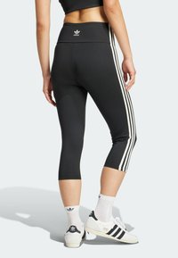Svart korta leggings med vita sidostrep, hög midja och Adidas-logotyp på baksidan. Kombineras med vita skor och strumpor.