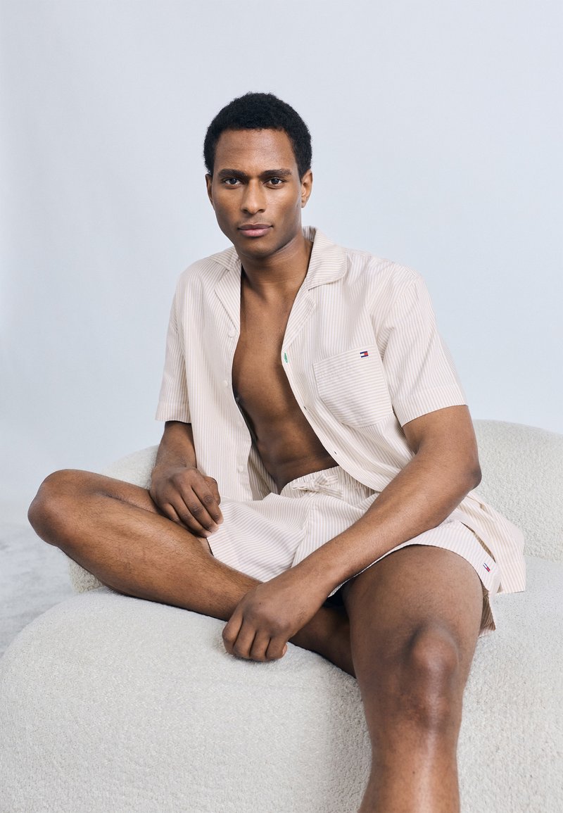 Φωτεινό μπεζ ριγωτό πουκάμισο με κοντά μανίκια και αντιστοιχισμένο σορτς από απαλό ύφασμα. Μικρή λεπτομέρεια λογότυπου στην τσέπη του πουκαμίσου, το μοντέλο κάθεται σε υφασμάτινη λευκή επιφάνεια.