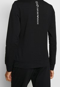 Svart sweatshirt med långa ärmar, ribbade muddar och nederkant. Har tryckt logotyptext vertikalt på baksidan. Mjuk, matt textur.