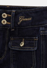 Sötétkék farmernadrág dupla gombos záródással, hímzett "Guess" márkanevével és dekoratív zsebbel fém díszekkel.