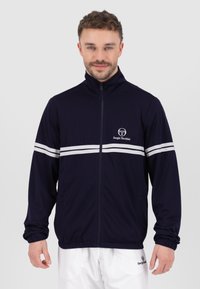Marinefarbene Sportjacke mit weißen horizontalen Streifen, Reißverschluss und strukturiertem Stoff. Mit Stehkragen und Logo auf der Brust.