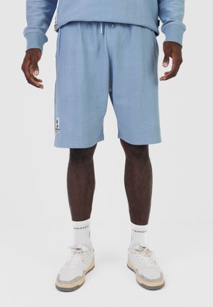 Mężczyzna w jasnoniebieskich prążkowanych szortach, białych skarpetkach z napisem oraz białych trampkach z beżowymi podeszwami, stojący na jednolitym tle.