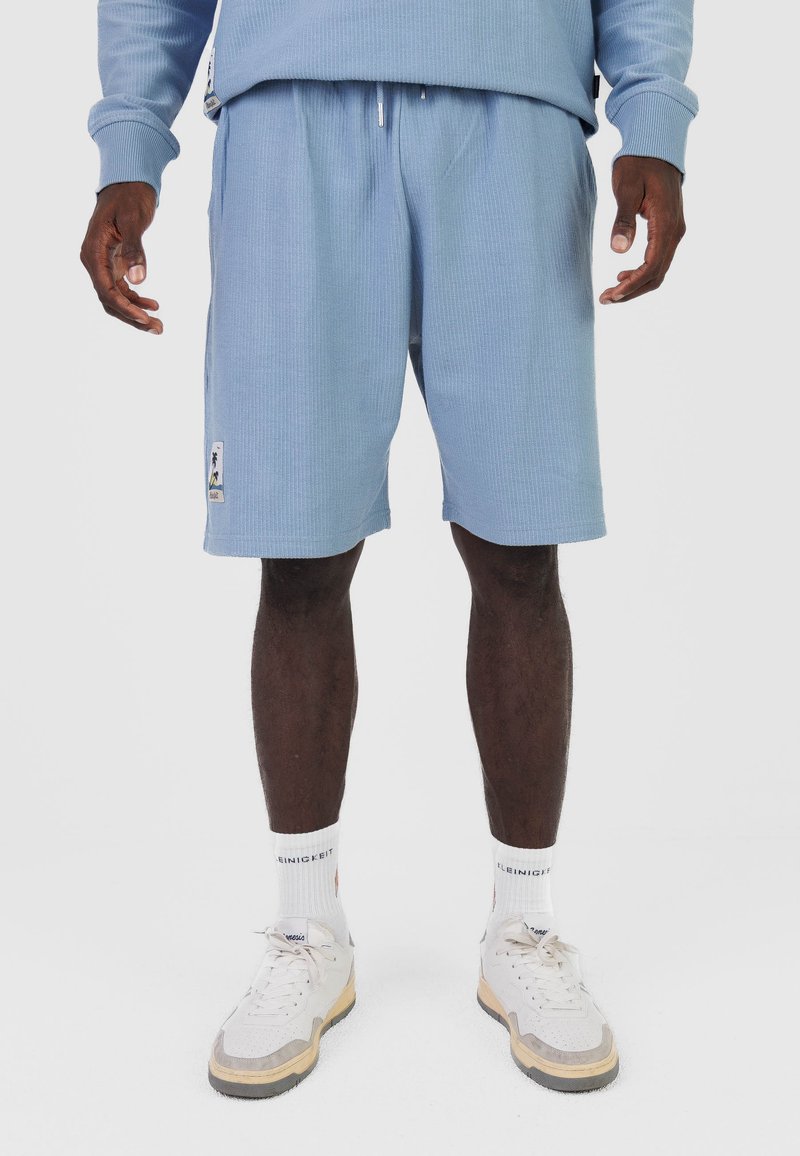 Homme portant un short côtelé bleu clair, des chaussettes blanches avec du texte et des baskets blanches avec des semelles beiges, debout devant un fond uni.